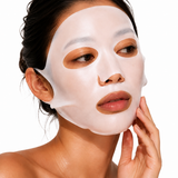A close-up of a woman wearing a white sheet mask on her face, with her hand touching her cheek, against a plain white background.